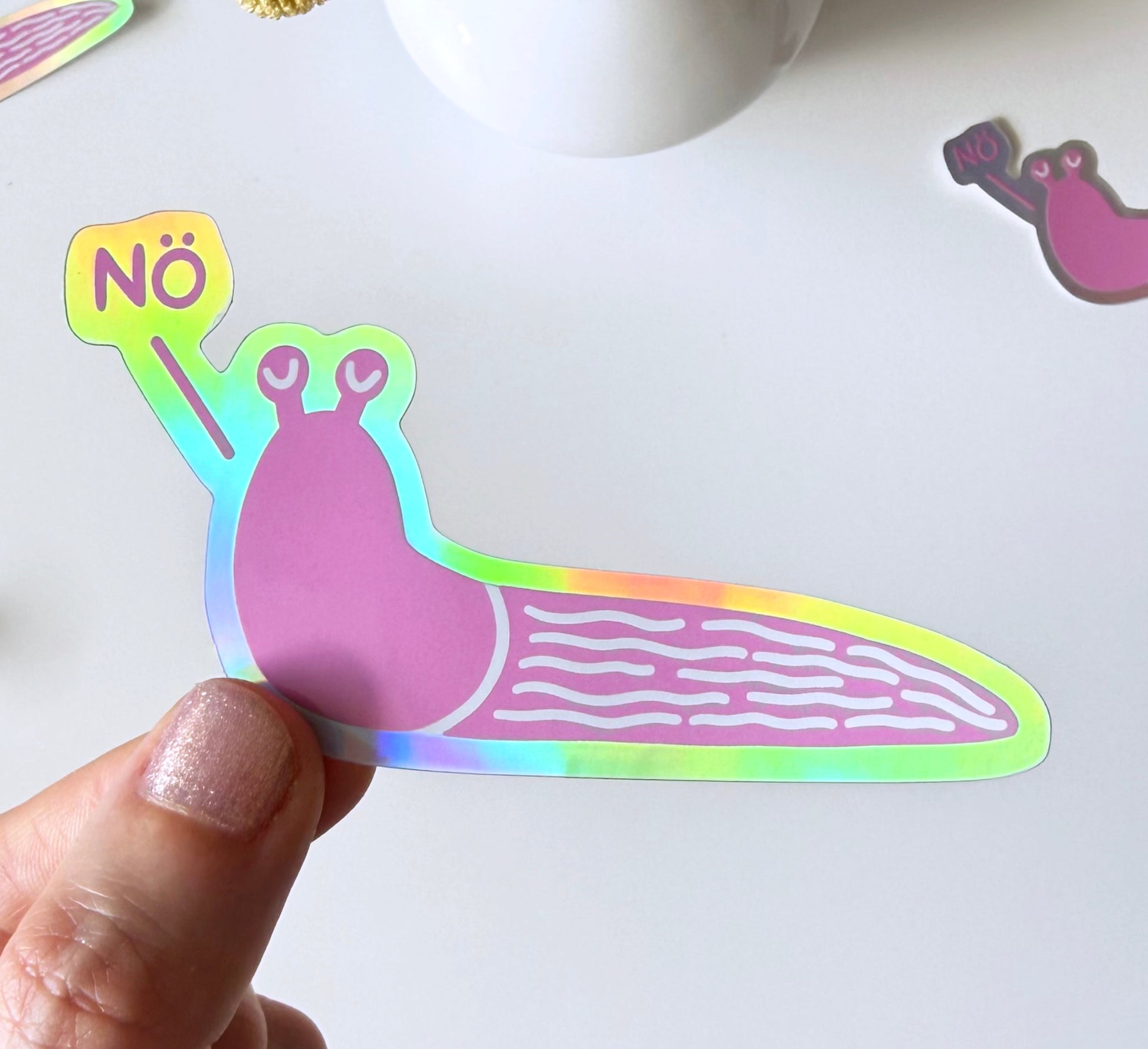A hand holds a die-cut sticker shaped like a pink snail with a holographic rainbow border and a small sign reading 'Nö' on a white surface.