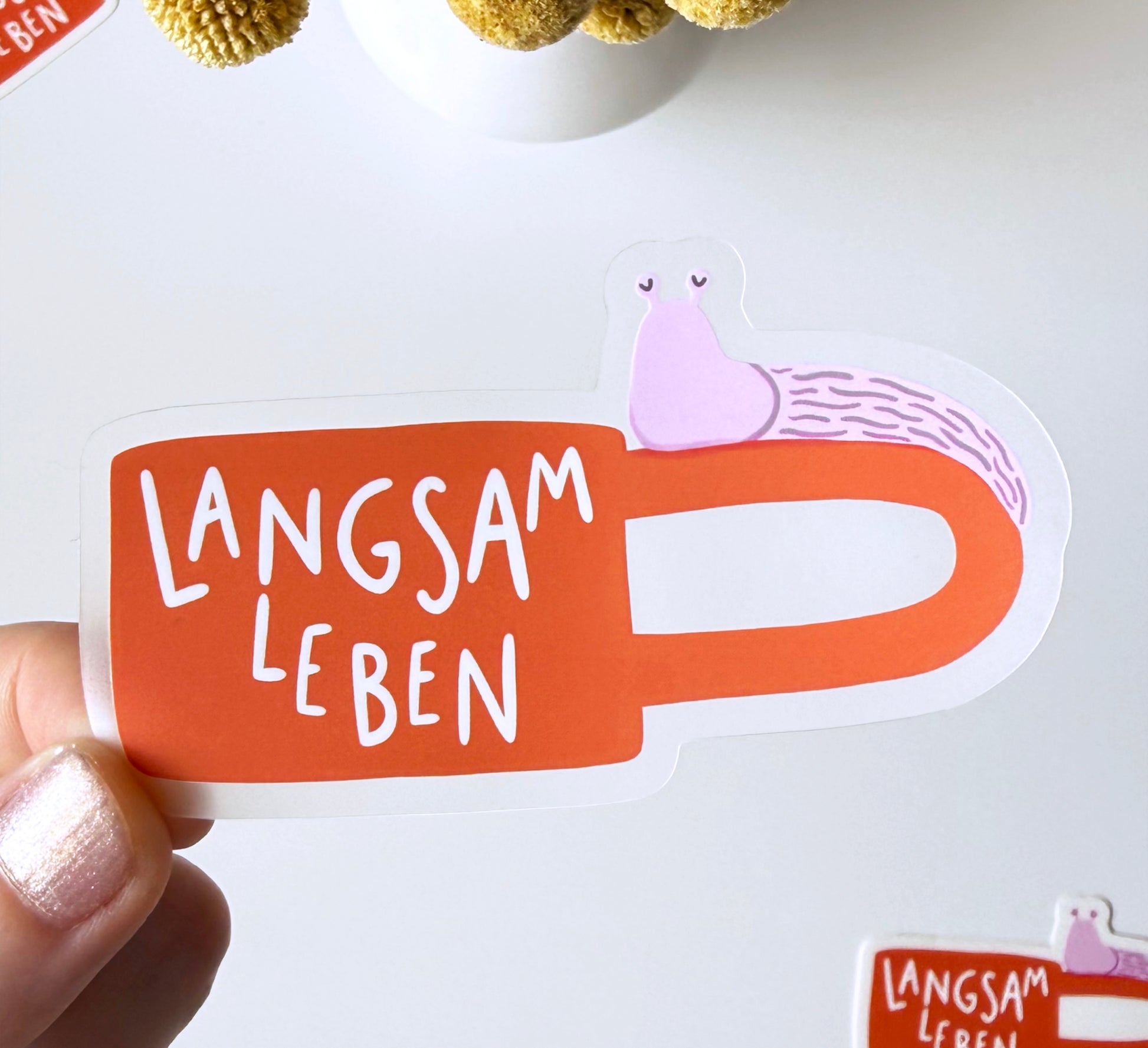 A hand holds a die-cut sticker shaped like an orange coffee mug with the German phrase 'LANGSAM LEBEN' in white lettering. A small pink snail is illustrated climbing up the side of the mug.