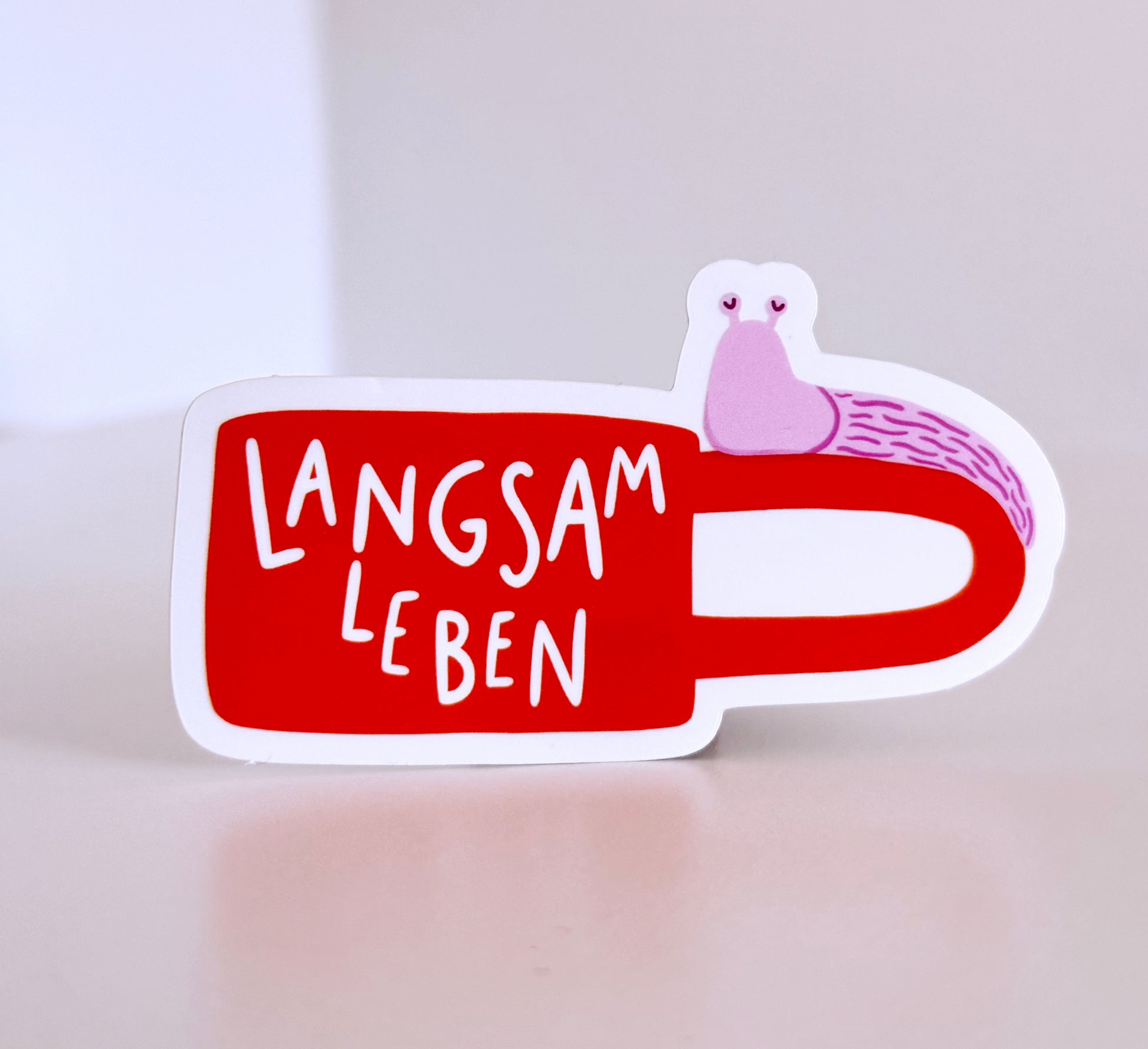 A die-cut sticker shaped like an orange coffee mug with the German phrase 'LANGSAM LEBEN' in white lettering and a small pink snail illustration on the handle, shown on a light beige surface against a neutral background.
